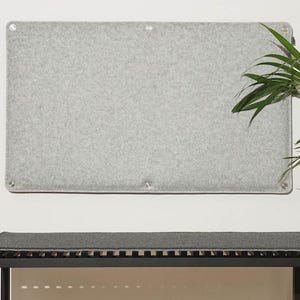 May include: A rectangular, light gray felt board mounted on a white wall. The board has metal grommets in each corner. Below, a dark gray bench with a slatted top and black metal frame is visible. Green plant leaves are in the background.