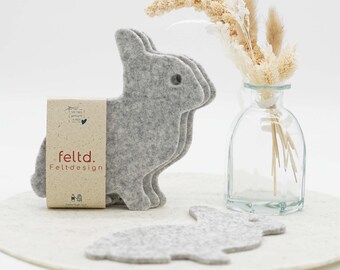 May include: A set of gray felt bunny-shaped coasters and a matching placemat. The coasters are stacked, and a single coaster lies flat. A small glass vase with dried flowers and the text "feldtd. Felt-design" are also visible.