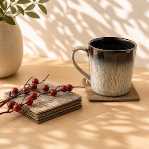 May include: A ceramic mug with a dark brown to white gradient sits on a square coaster. A stack of square coasters and a sprig of red berries are also on the wooden surface. A beige vase with green leaves is in the background.