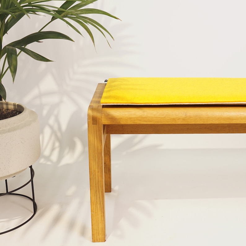 Puede incluir: Un banco de madera con un coj&iacute;n amarillo brillante. El banco tiene un asiento rectangular y cuatro patas rectas. Una planta en maceta con hojas verdes est&aacute; a la izquierda del banco.