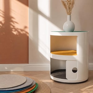 Puede incluir: Una moderna casa para gatos blanca con un diseño redondeado. Cuenta con una parte superior abierta, un estante central con un cojín amarillo y un compartimento inferior con una puerta. Una pequeña vasija blanca con flores secas encima.