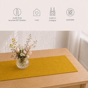 Könnte beinhalten: Ein senfgelber Tischläufer auf einem hellen Holztisch mit einer kleinen Glasvase mit Blumen. Der Läufer ist rechteckig und aus filzartigem Material. Text im Bild: aus recycelten PET-Flaschen, gemütlich, hergestellt in Köln, waschbar.