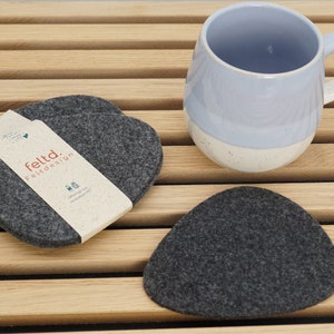 Op de afbeelding: Een set grijze vilten onderzetters en een lichtblauwe en witte mok op een houten oppervlak. De onderzetters zijn hartvormig en driehoekig. De verpakking leest "feltd. Feltdesign."