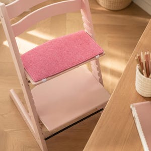 May include: A pink wooden high chair with a pink felt seat and footrest. The chair has a curved back and adjustable features. The chair is next to a wooden table with pencils and a notebook.