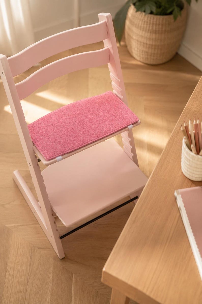 May include: A pink wooden high chair with a pink felt seat and footrest. The chair has a curved back and adjustable features. The chair is next to a wooden table with pencils and a notebook.