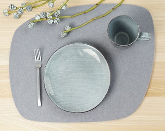 Op de afbeelding: Een lichtgrijze, organisch gevormde placemat van vilt met een lichtblauw keramisch bord met witte horizontale lijnen, een bijpassende keramische mok en een roestvrijstalen vork. Erboven ligt een decoratieve tak met kleine groene knoppen.