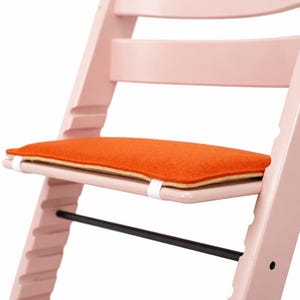 May include: A light pink wooden high chair with an orange seat cushion. The high chair has a black footrest and white straps securing the cushion. The design is modern and minimalist.