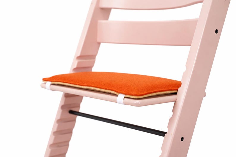 May include: A light pink wooden high chair with an orange seat cushion. The high chair has a black footrest and white straps securing the cushion. The design is modern and minimalist.