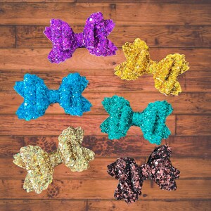 Könnte beinhalten: Sechs glitzernde Haarbänder in verschiedenen Farben, darunter Lila, Türkis, Gold und Braun. Die Schleifen sind auf einer Holzoberfläche angeordnet.