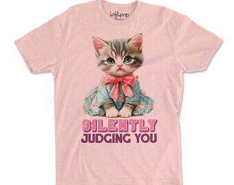 May include: Pink t-shirt featuring a graphic of a kitten wearing a blue dress with a pink bow. The text "SILENTLY JUDGING YOU" is printed below the kitten. The shirt has a crew neck and short sleeves.