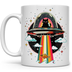 May include: A white ceramic mug with a black cat sitting in a flying saucer. The saucer is flying through a rainbow with a fish swimming below. The background is a dark blue with stars.