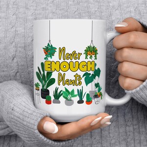 Puede incluir: Taza de cer&aacute;mica blanca con un dise&ntilde;o amarillo y verde. La taza presenta una variedad de plantas en maceta y el texto "Never Enough Plants".