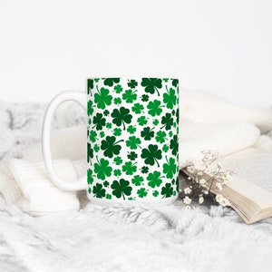 May include: White ceramic mug with a green shamrock pattern. The mug has a large handle and is sitting on a white fuzzy surface.
