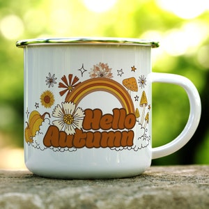 Pu&ograve; includere: Una tazza di smalto bianco con un design a arcobaleno, funghi e fiori. La tazza dice "Hello Autumn" in lettere marroni.