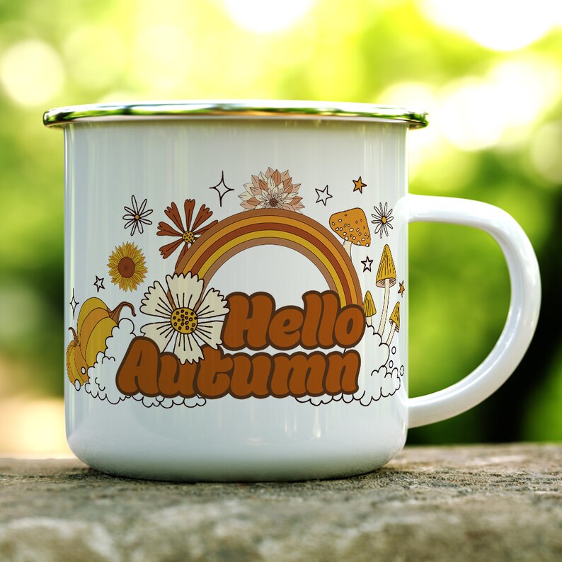 Pu&ograve; includere: Una tazza di smalto bianco con un design a arcobaleno, funghi e fiori. La tazza dice "Hello Autumn" in lettere marroni.