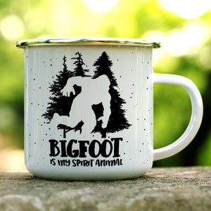 May include: White enamel camping mug with a black silhouette of Bigfoot walking through a forest. The text "BIGFOOT is my spirit animal" is printed below the image.