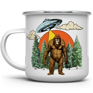 May include: White enamel mug featuring a Bigfoot illustration under a UFO with a forest and sunset backdrop. The design includes a brown Bigfoot, a blue UFO, and green trees. The mug has a white handle and a silver rim.