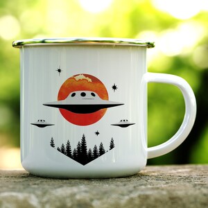 May include: White enamel mug with a red, orange and yellow sunset design. The mug features a black silhouette of trees and three flying saucers. The mug has a silver rim and handle.