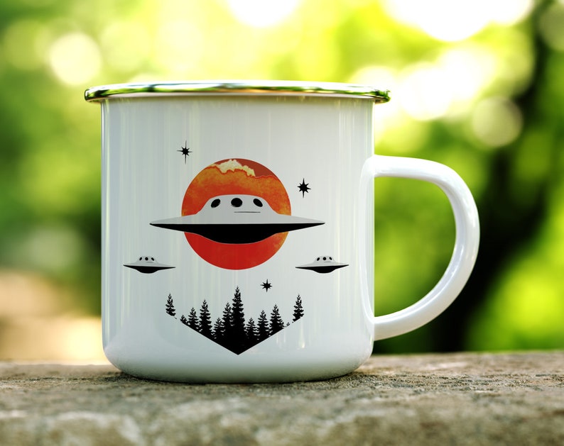 May include: White enamel mug with a red, orange and yellow sunset design. The mug features a black silhouette of trees and three flying saucers. The mug has a silver rim and handle.