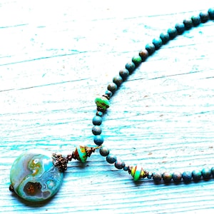 以下が含まれることがあります： 青、緑、茶色の渦巻き模様が特徴の、大きくて丸いペンダントが付いた手作りのネックレス。 ネックレスは、小さくて丸い青緑色のビーズのストランドで、緑、茶色、黄色の装飾ビーズでアクセントが付けられています。