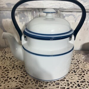 May include: A white ceramic teapot featuring a smooth finish with thin navy blue horizontal bands around the body and lid. The teapot includes a curved spout and a tall, arched black handle that extends over the top of the lid.