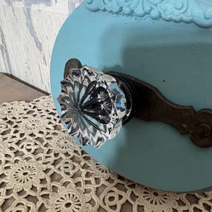 May include: A light blue circular wooden plaque featuring a decorative scrollwork top and a clear, faceted glass knob mounted on a dark, weathered metal plate. The object has a smooth, matte finish and a distinct, multi-faceted crystal design.