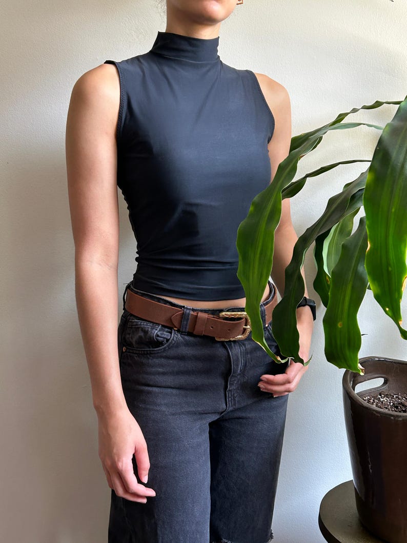 Peut inclure: Un haut noir sans manches &agrave; col montant, associ&eacute; &agrave; un short en jean noir et une ceinture en cuir marron avec une boucle dor&eacute;e. Un pot de plante est visible &agrave; droite.