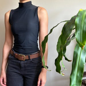 Peut inclure: Un haut noir sans manches &agrave; col montant, associ&eacute; &agrave; un jean en denim noir et une ceinture en cuir marron avec une boucle dor&eacute;e. Une plante verte &agrave; longues feuilles est &agrave; droite. La ceinture mesure environ 3,8 cm de large.