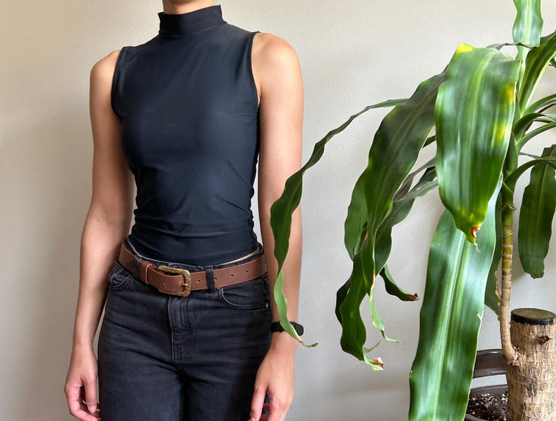 Peut inclure: Un haut noir sans manches &agrave; col montant, associ&eacute; &agrave; un jean en denim noir et une ceinture en cuir marron avec une boucle dor&eacute;e. Une plante verte &agrave; longues feuilles est &agrave; droite. La ceinture mesure environ 3,8 cm de large.