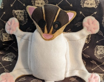 May include: A white plush toy shaped like a sugar glider with brown ears and a brown stripe down its back. The toy has pink paw pads and a long white tail.