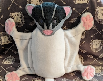 May include: A plush sugar glider toy in white, gray, black, and pink. The toy has outstretched wings and a long tail. The background is a gray fabric with a pattern of hedgehogs and arrows.