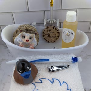 May include: A white miniature bathtub containing a hedgehog figurine, a round wooden sign with the text Sew Magick, and a bottle labeled Quill Cleanser. A white towel with a blue hedgehog outline, a toothbrush, and nail clippers are in front.