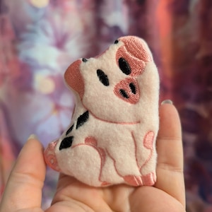 Op de afbeelding: Een klein roze pluche varken met zwarte geborduurde ogen en details. Het varken zit met zijn poten onder zich.