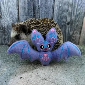 May include: A purple plush bat with pink and blue embroidery. The bat has large, round eyes and a heart-shaped nose. The bat is sitting on a gray surface.