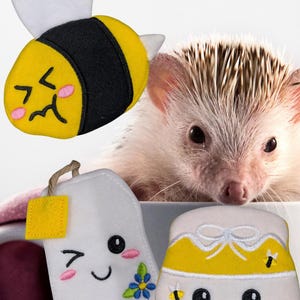 May include: A felt bee, tea bag, and honey pot with smiling faces and a hedgehog. The bee is yellow and black with white wings. The tea bag is white with a yellow tag and blue flower. The honey pot is yellow and white.