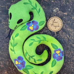 May include: A green felt snake with embroidered vines and purple flowers. The snake is curled into a spiral shape. The text "Sew Magick" is on a brown tag.