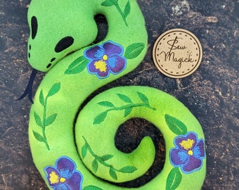 Könnte beinhalten: Eine grüne Filzschlange mit gestickten Ranken und lila Blüten. Die Schlange ist zu einer Spirale zusammengerollt. Der Text "Sew Magick" befindet sich auf einem braunen Etikett.
