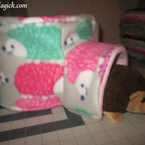 May include: A plush hedgehog hideaway with a tunnel entrance. The hideaway is made of soft, gray fleece with pink and green hedgehog patterns. The tunnel entrance is lined with pink fleece. A brown plush hedgehog is peeking out of the tunnel entrance.