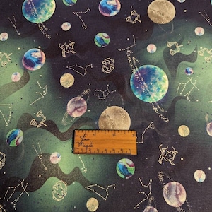 May include: A dark blue fabric with a repeating pattern of planets, stars, and constellations in gold and green. The fabric has a subtle gradient from dark green to black. A ruler with the words "Sew Magick" is included in the image.