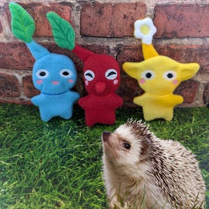 May include: Three plush toys, a blue one with green leaves, a red one with a beak, and a yellow one with a white flower on top, are sitting on a green grassy surface. A small hedgehog is looking at the toys.