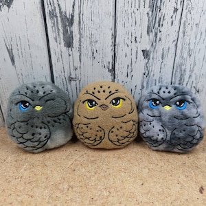 May include: Three plush owl toys with embroidered faces. The owls are gray, brown, and a camouflage pattern. Each owl has yellow and blue eyes.