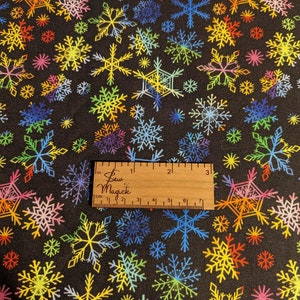 May include: Black fabric with a repeating pattern of colorful snowflakes. The fabric is measured with a ruler that reads "Sew Magick" in inches and centimeters.