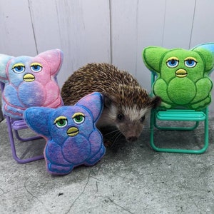 May include: A small hedgehog sits in front of three plush toys. The toys are shaped like a fuzzy, colorful creature with large eyes and a beak. The toys are sitting on small folding chairs. The chairs are purple, green, and blue.