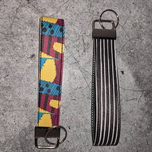 Könnte beinhalten: Zwei Schlüsselanhänger, einer mit einem bunten geometrischen Muster und der andere mit schwarzen und weißen Streifen. Beide Schlüsselanhänger haben silberne Schlüsselringe.