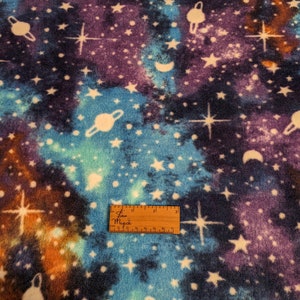 May include: A blue, purple, and orange fabric with a white galaxy print. The print features stars, planets, and moons. The fabric is soft and fuzzy, perfect for making blankets, pillows, or other crafts.