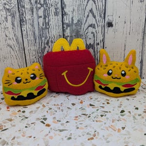 Pode incluir: Três brinquedos de pelúcia de feltro consistindo em uma caixa vermelha com um logotipo de arco amarelo e um sorriso amarelo, ladeada por dois personagens de gato em forma de hambúrguer amarelo com características faciais bordadas e detalhes.