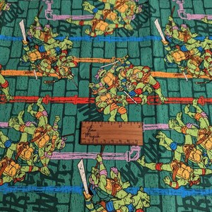 May include: Green fabric with a repeating pattern of Teenage Mutant Ninja Turtles characters. The turtles are in various poses, some are fighting, some are standing, and some are jumping. The fabric has a brick wall background with the words "Cowabunga" and "Teenage Mutant Ninja Turtles" written in white.