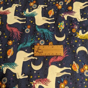 May include: A blue fabric with a repeating pattern of white unicorns, colorful planets, stars, and flowers. The fabric has a textured surface and a ruler is included for scale.