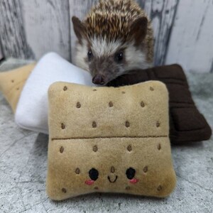 Op de afbeelding: Een bruin en wit  egeltje kijkt naar een pluche s'more. De s'more is gemaakt van drie pluche stukken: een witte marshmallow, een bruine graham cracker en een bruine chocolade reep.