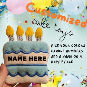May include: A blue and white felt cake toy with yellow candles and the text "NAME HERE". The toy is customizable with different colors, candle numbers, and a name or happy face.
